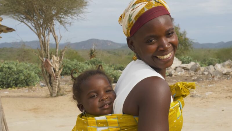 A Smiling Woman Wearing a Headscarf Carries a Young Child on Her Back, Surrounded by A Rural Landscape with Trees and Mountains in The Background, Highlighting the Natural Diversity and Cultural Warmth of West Africa